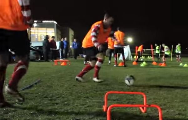 A football player dribbles a ball near orange cones and agility hurdles at night on a grassy field. Other players and spectators are visible in the background.