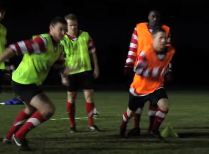 A football player sprints on a field at night; he wears a yellow and red-and-white striped shirt, while others around him wear similar uniforms.