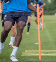 A football player runs past orange and yellow agility cones during a training session on a grassy field.