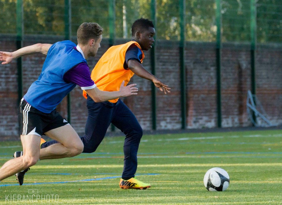 A football player, wearing an orange bib, runs, dribbling; another player in blue pursues; the context is a grassy football field.